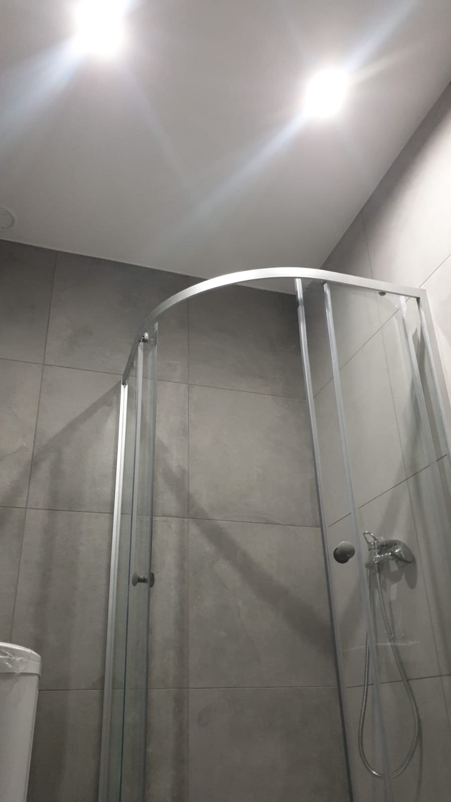 Szklana kabina prysznicowa z chromowanymi profilami na tle szarych, kwadratowych płytek w minimalistycznej łazience z widocznym fragmentem toalety.