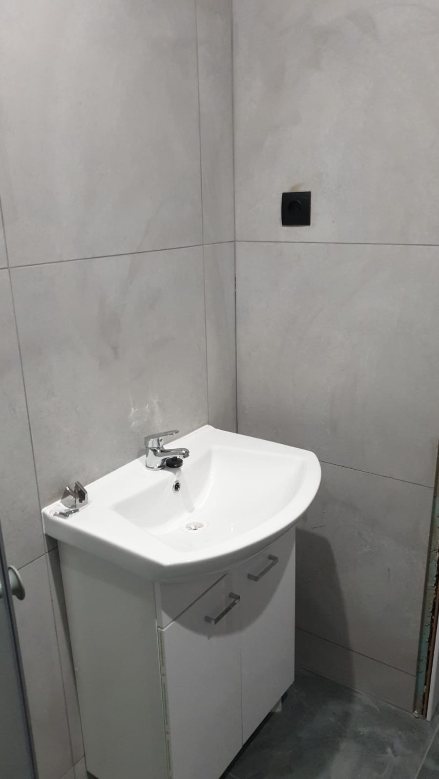 Biała umywalka narożna z szafką w łazience z szarymi płytkami, widoczny czarny włącznik na ścianie i akcesoria montażowe na umywalce.