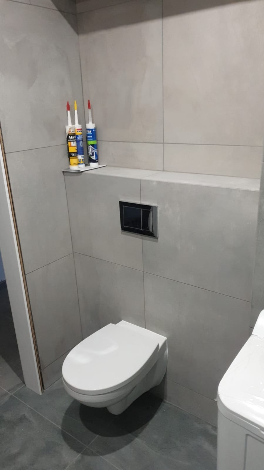 Wisząca toaleta z czarnym przyciskiem spłukującym na tle szarych płytek, z trzema tubami silikonu na półce powyżej.