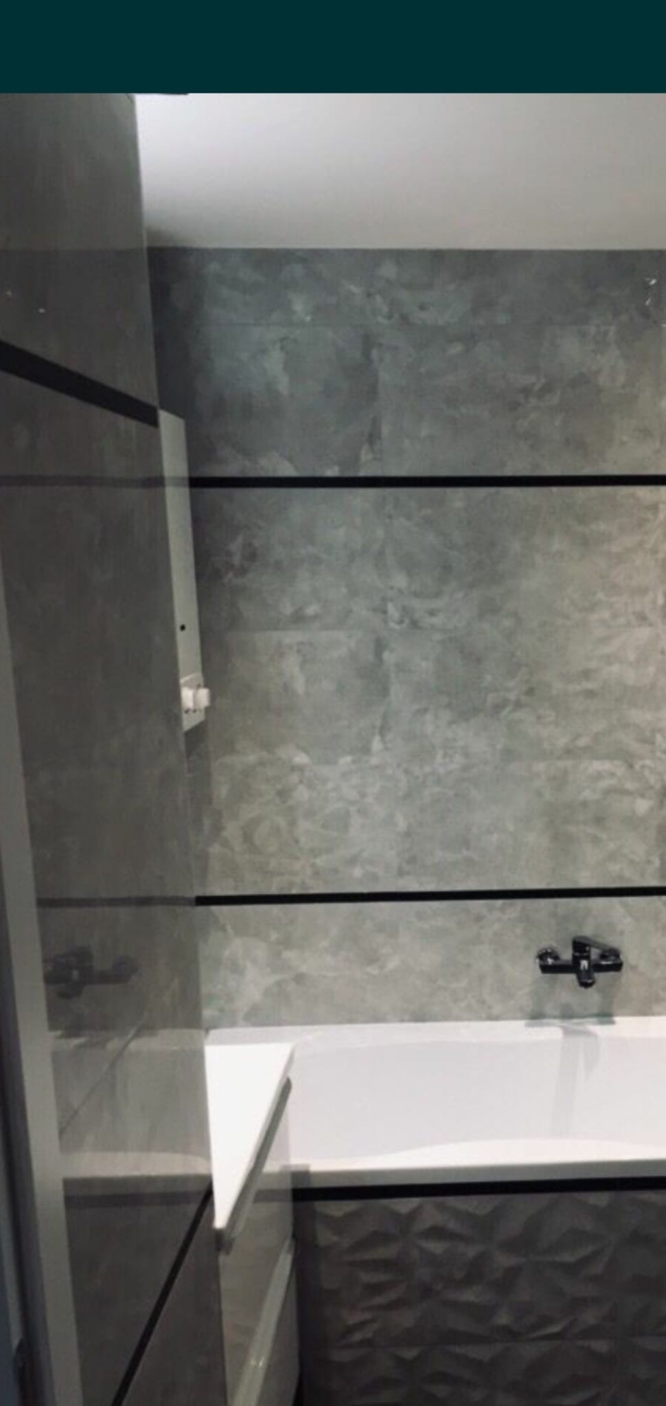 Wykończona łazienka z szarymi płytkami imitującymi kamień, czarnymi listwami dekoracyjnymi, białą wanną i umywalką z szafką o geometrycznym wzorze.