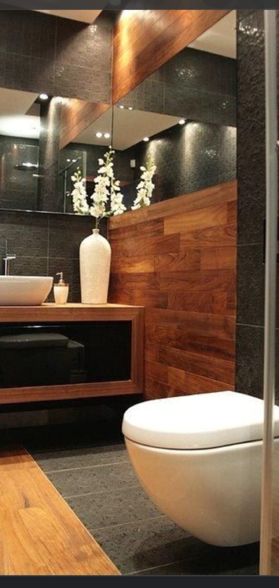 Nowoczesna łazienka z ciemnymi płytkami, drewnianymi panelami i wiszącą toaletą, oświetlona punktowymi lampami, z białym umywalkiem na drewnianym blacie i wazonem z białymi kwiatami.