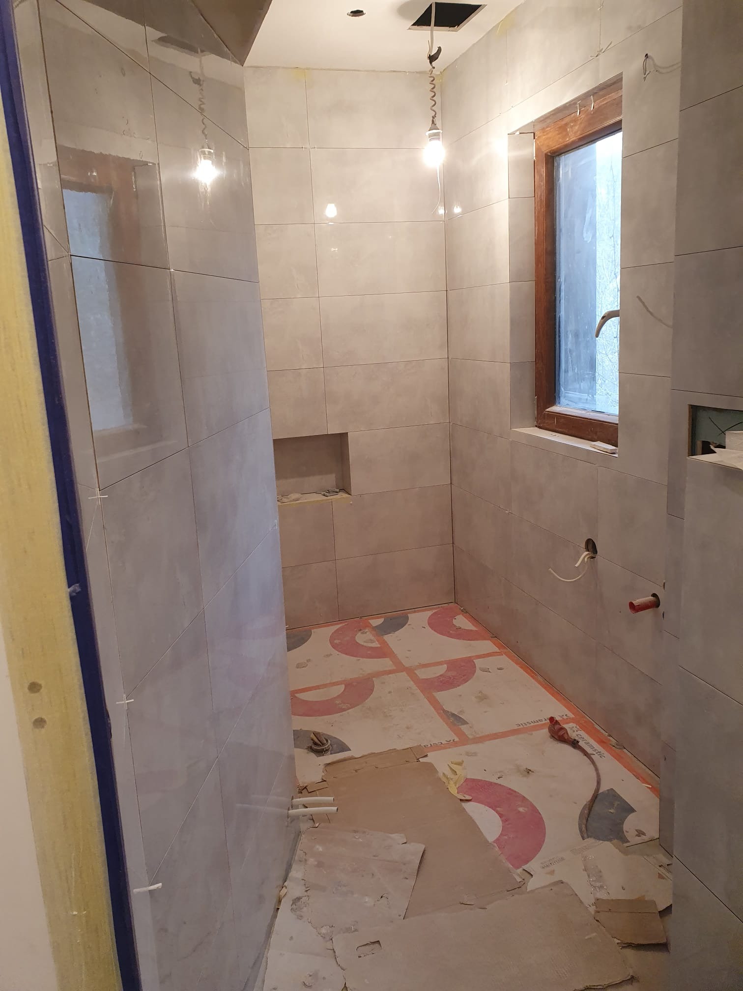 Ściany łazienki w trakcie układania płytek, widoczne krzyżyki dystansowe, przygotowane otwory na instalacje, zabezpieczona ściana z oknem, podłoga częściowo zakryta płytami ochronnymi.