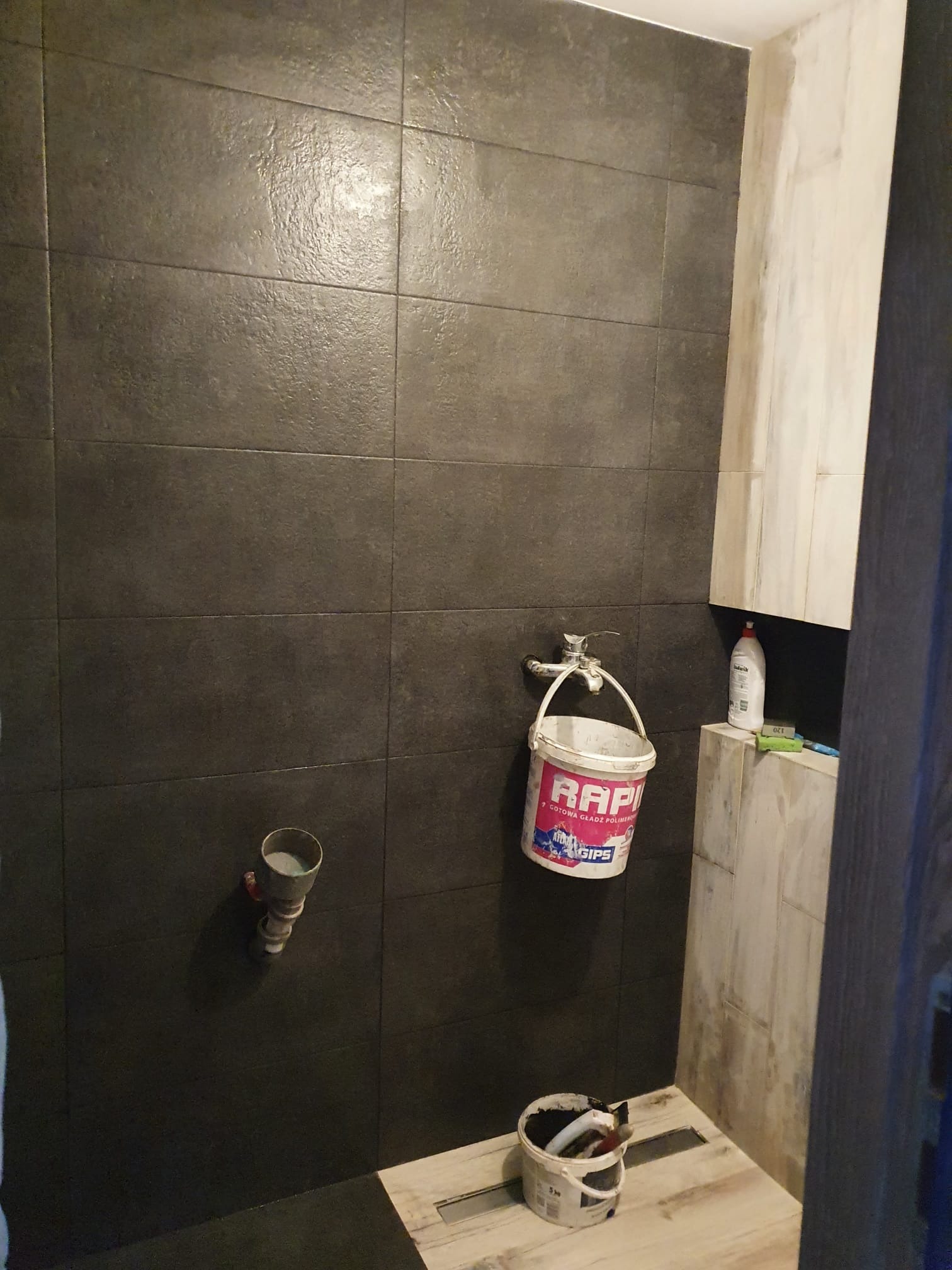 Ciemne, kwadratowe płytki na ścianie w trakcie prac wykończeniowych w łazience, widoczne wiaderka po gładzi i rura wystająca ze ściany.