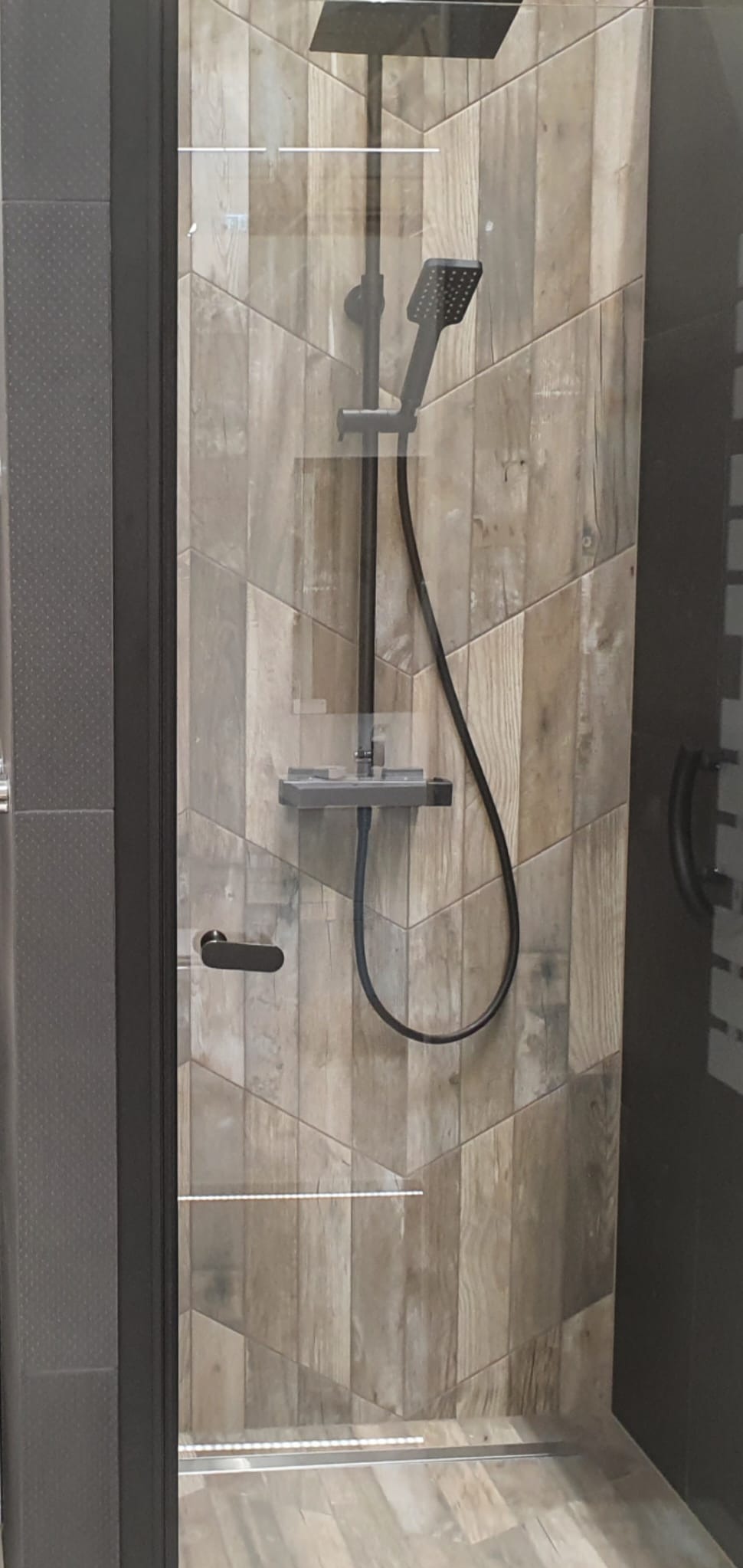 Kabina prysznicowa z deszczownicą w kolorze czarnym, ściany wykończone płytkami imitującymi drewno ułożonymi w jodełkę.