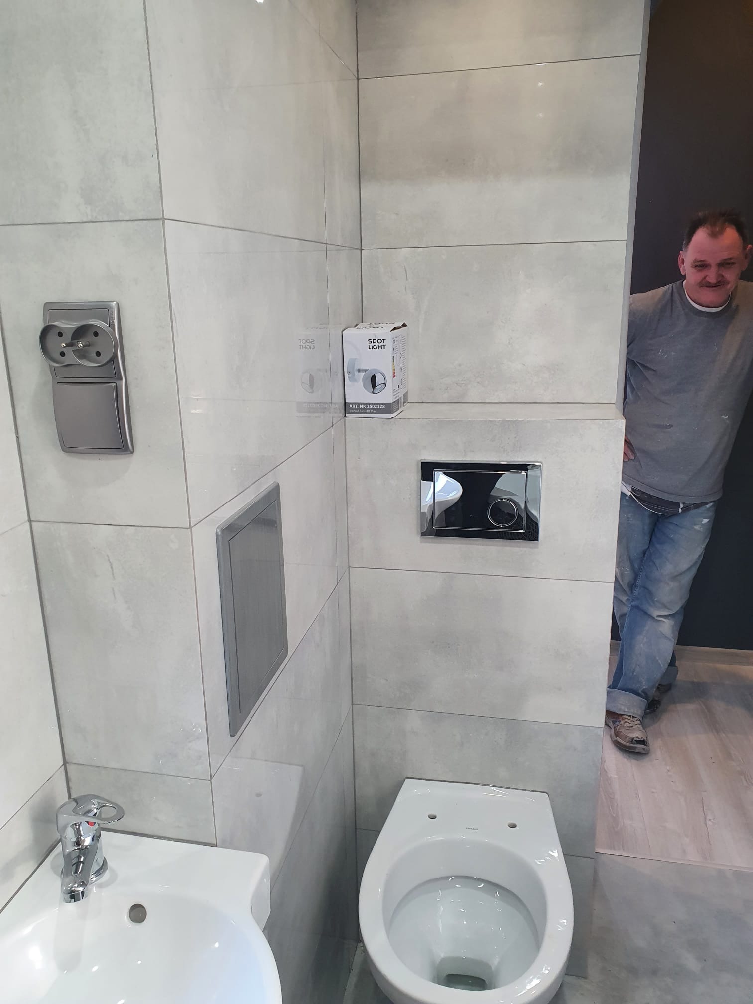 Wykończona łazienka z szarymi płytkami, białą umywalką i toaletą, widoczny pracownik w tle.