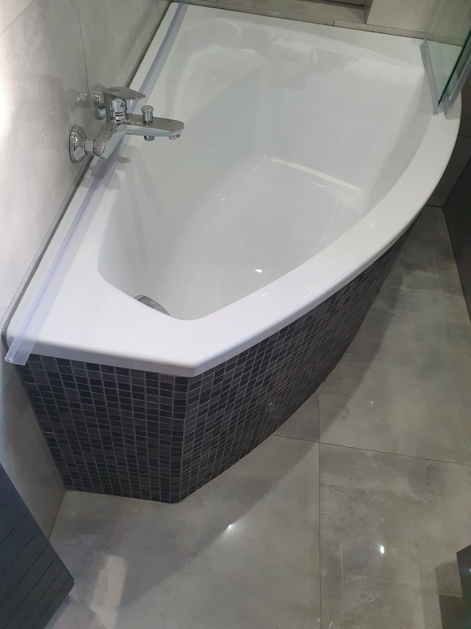 Narożna wanna z obudową wykończoną mozaiką w odcieniach szarości, bateria chromowana, jasne płytki podłogowe i ścienne.