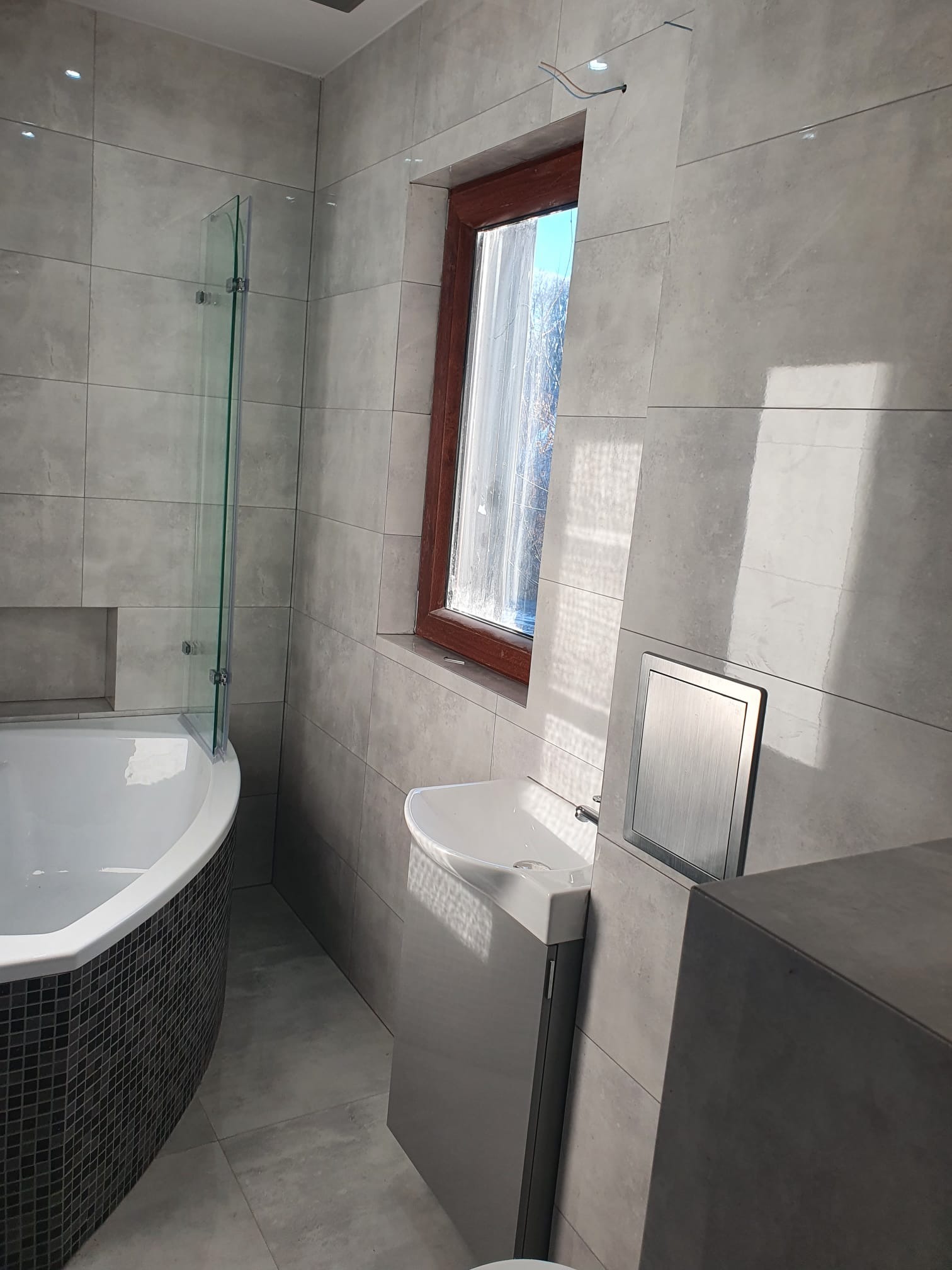 Nowoczesna łazienka z szarymi płytkami, owalną wanną z mozaiką, wiszącą umywalką i oknem w drewnianej ramie. Widoczny panel dostępowy do instalacji.