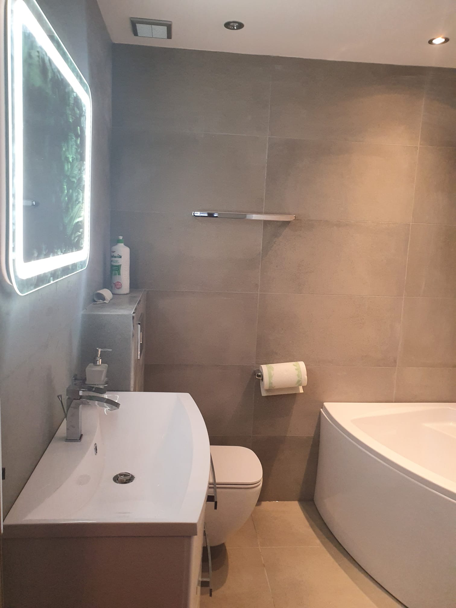 Nowoczesna łazienka z płytkami w odcieniach szarości, podświetlane lustro, biała umywalka z szafką, toaleta i narożna wanna.