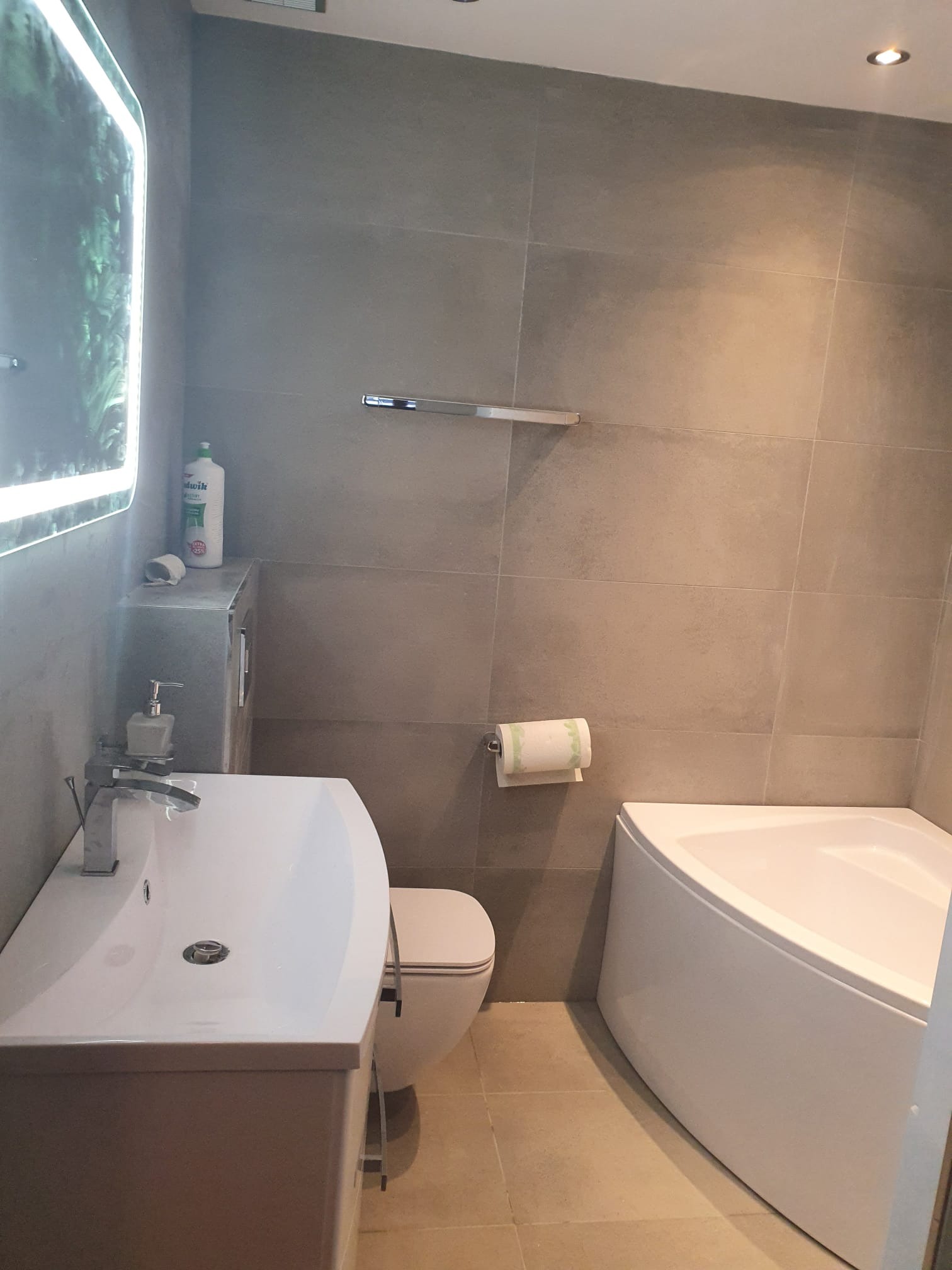 Nowoczesna łazienka z narożną wanną, umywalką z szafką, toaletą i dużymi szarymi płytkami na ścianach, oświetlona lampą LED w lustrze.