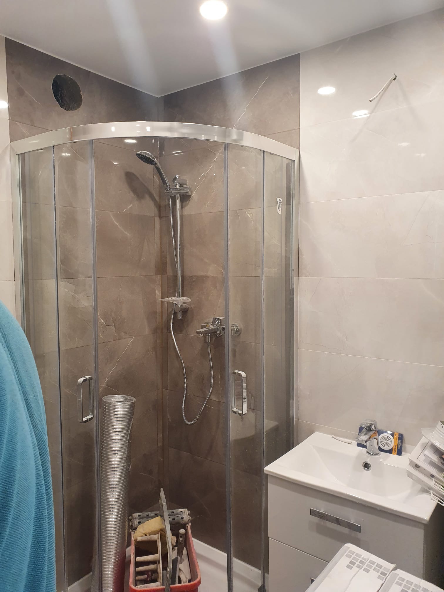 Niedokończona łazienka z kabiną prysznicową narożną, widoczny otwór w ścianie, narzędzia w brodziku, umywalka z baterią i materiały wykończeniowe obok.