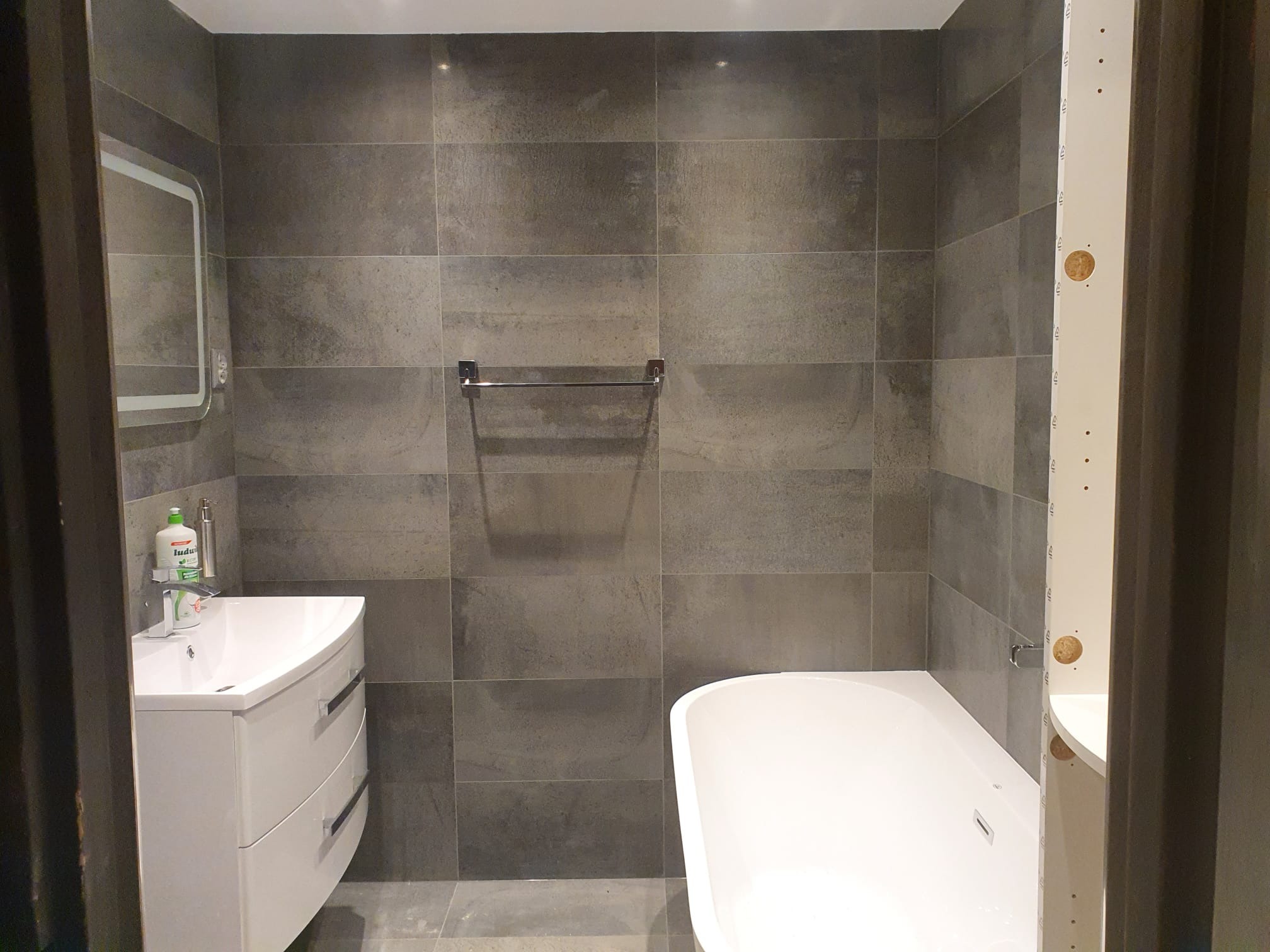 Wykończona łazienka z szarymi płytkami imitującymi kamień, białą umywalką z szafką i wanną, widoczny fragment ramy drzwiowej z otworami montażowymi.