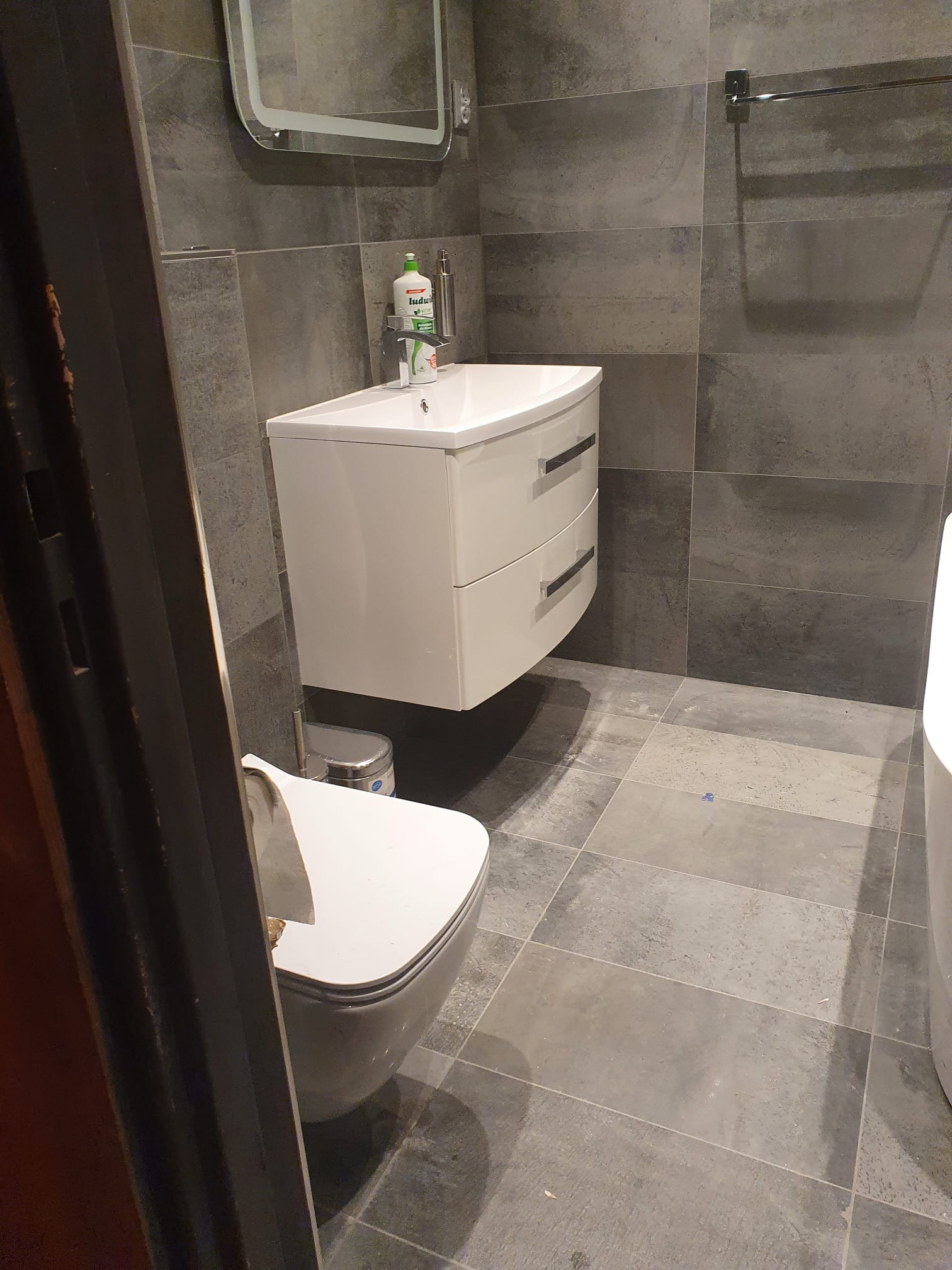 Nowoczesna łazienka z szarymi płytkami, białą wiszącą szafką z umywalką, chromowaną baterią, okrągłym lustrem z podświetleniem LED i wiszącą toaletą.