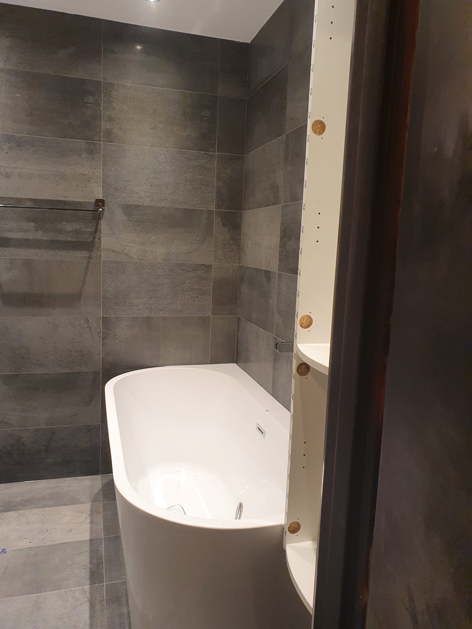 Wanna wolnostojąca w łazience z szarymi płytkami, widoczny fragment montowanej szafki z półkami i kołkami mocującymi.