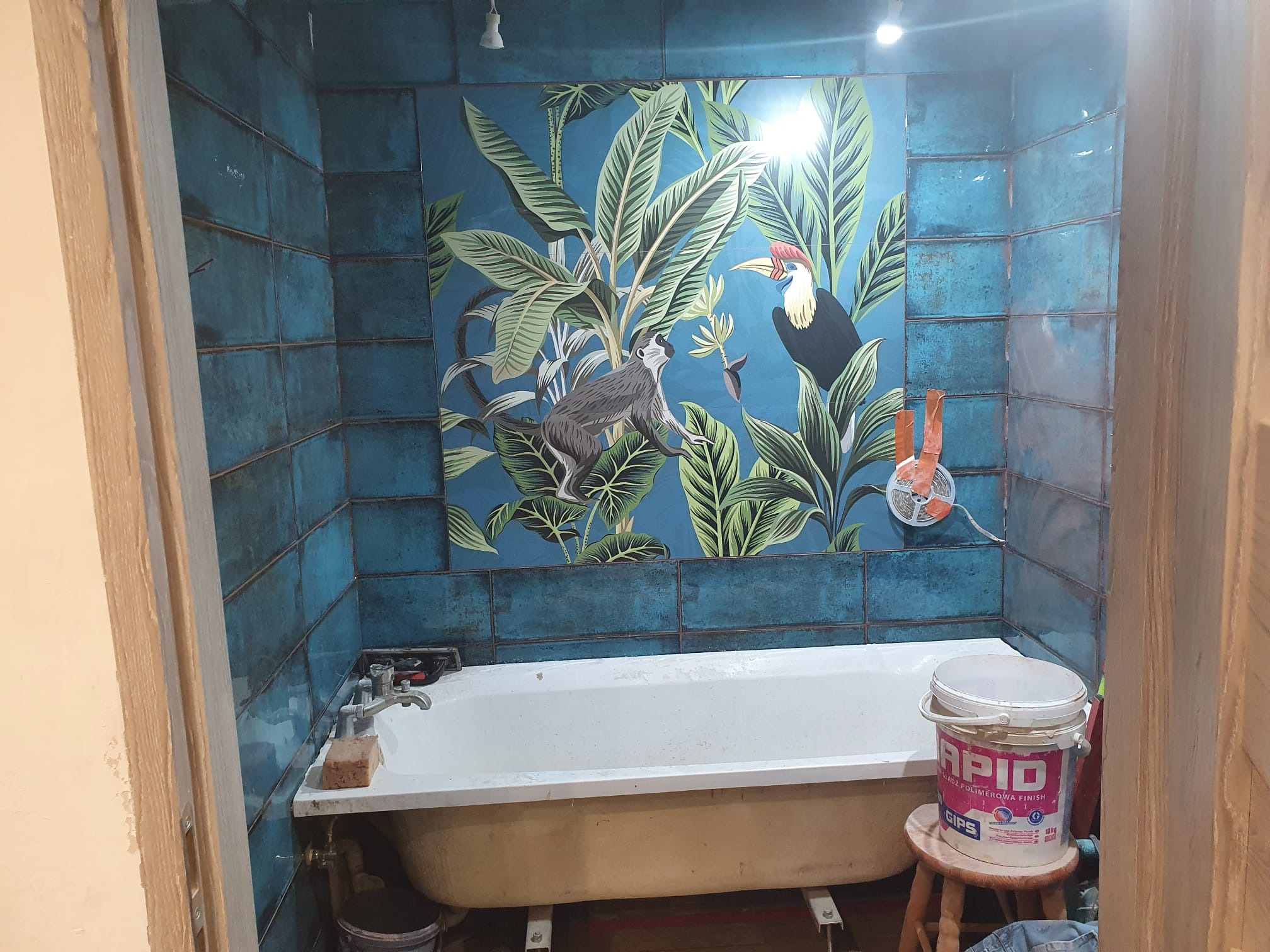 Niebieskie płytki imitujące patynę z motywem tropikalnym na ścianie nad wanną, widoczny kubeł z gładzią polimerową na stołku.