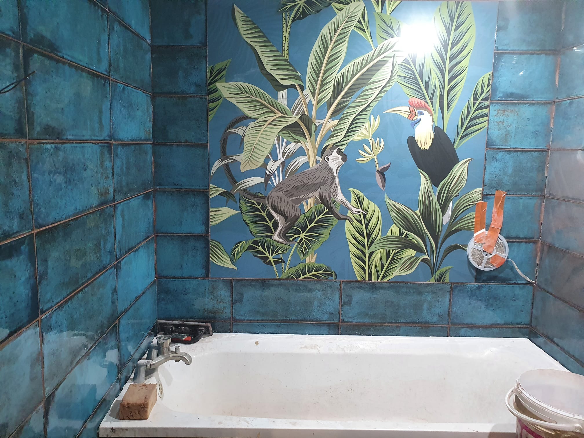 Turkusowe płytki w łazience z motywem dżungli na ścianie, widoczna wanna i akcesoria montażowe.