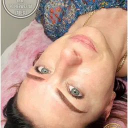 Kobieta leżąca na różowym podkładzie z widocznymi brwiami po zabiegu permanentnym, nałożony czepek ochronny, widoczne logo firmy w prawym dolnym rogu.