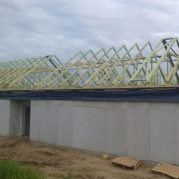 Konstrukcja drewniana dachu w trakcie budowy, widoczne elementy więźby dachowej na tle szarej ściany budynku i pochmurnego nieba. Częściowo zabezpieczona czarną folią.