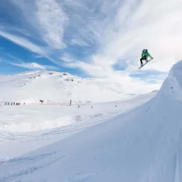 Snowboardzista w zielonej kurtce skacze na tle zaśnieżonych gór i błękitnego nieba z chmurami.