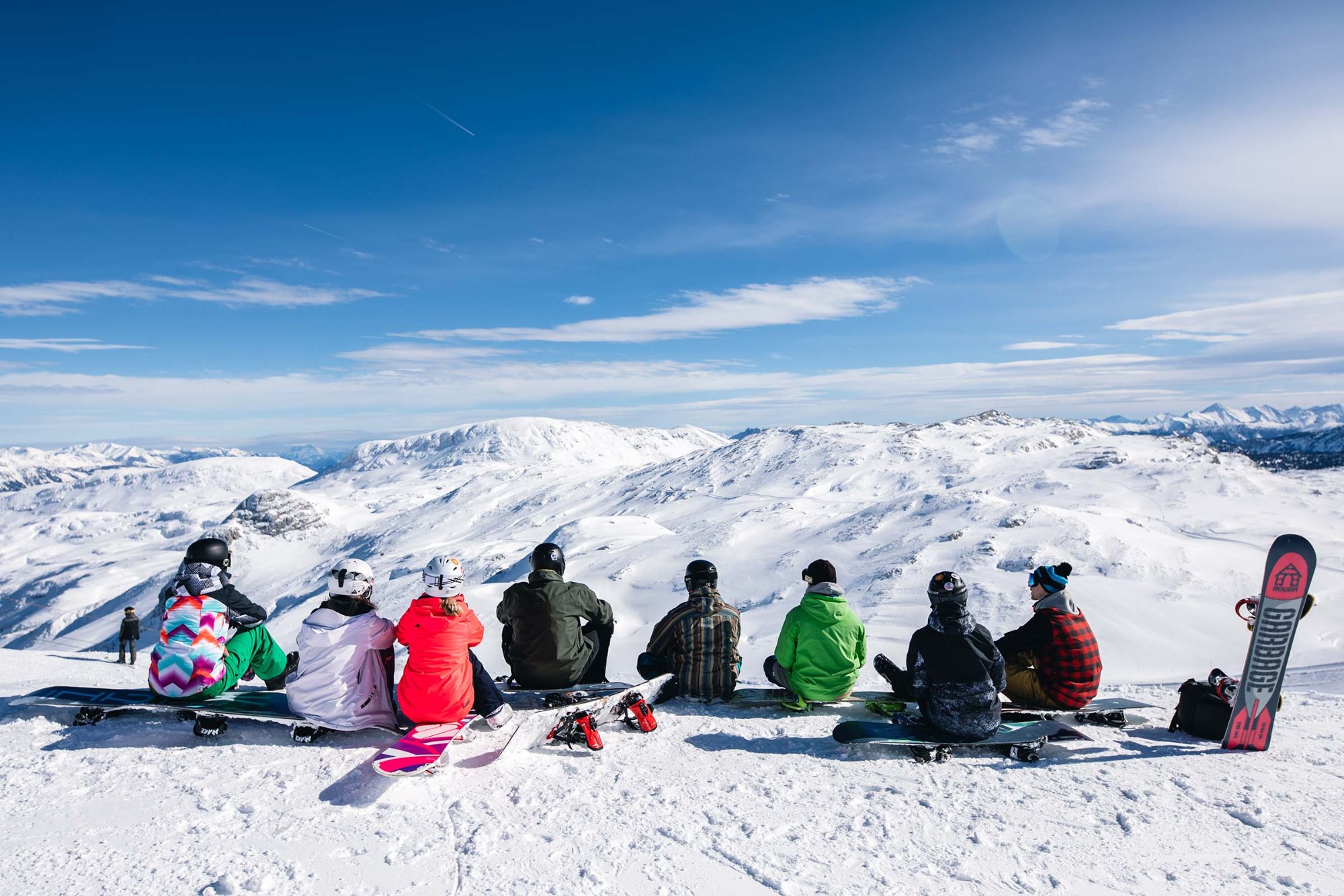 Grupa snowboardzistów siedzi na deskach, podziwiając widok na ośnieżone góry i błękitne niebo z smugami kondensacyjnymi samolotów.
