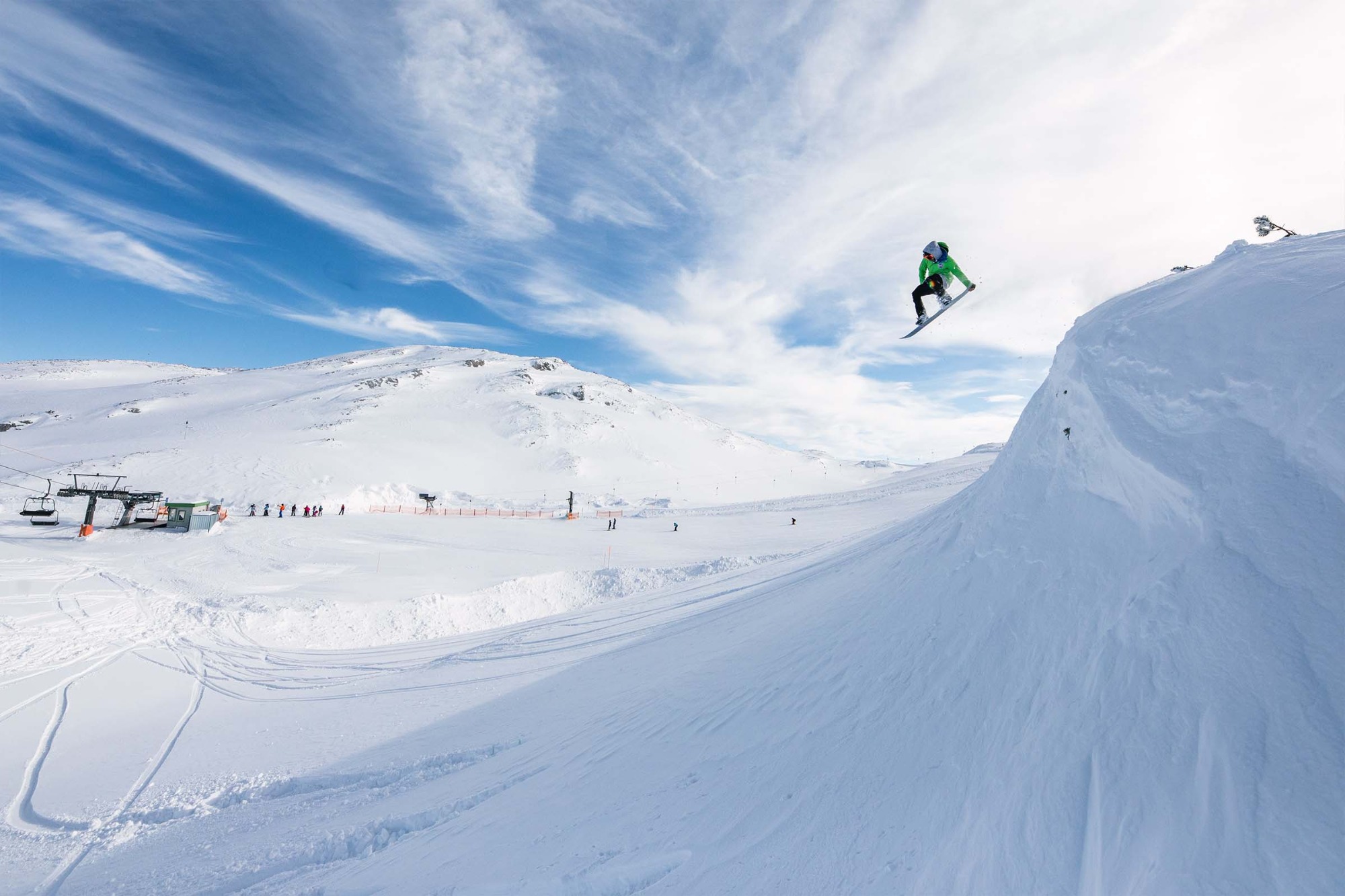 Snowboardzista w zielonej kurtce skacze na tle zaśnieżonych gór i błękitnego nieba z chmurami.