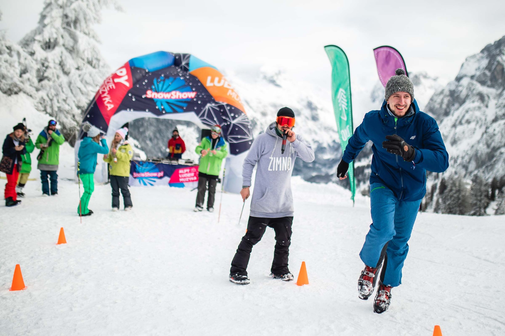 Zawody na śniegu: mężczyzna w butach narciarskich biegnie slalomem między pomarańczowymi pachołkami, w tle scena z DJ-em i publicznością podczas wydarzenia SnowShow.