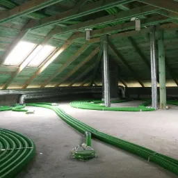 Montaż instalacji rekuperacji w poddaszu: zielone, karbowane rury wentylacyjne rozłożone na betonowej podłodze, widoczne elementy mocujące, drewniana konstrukcja dachu z oknem połaciowym.