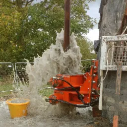 Wiercenie otworu: pomarańczowa maszyna w akcji, tryskająca woda i błoto, żółte wiadro zbierające płyny, drzewa w tle.