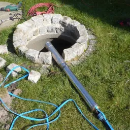 Montaż pompy głębinowej w studni z kamienną obudową, widoczne narzędzia i kable na trawie.