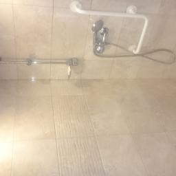 Piotr Grochola - Jasna łazienka z beżowymi płytkami, zamontowany prysznic z uchwytem dla niepełnosprawnych oraz składane krzesełko prysznicowe.