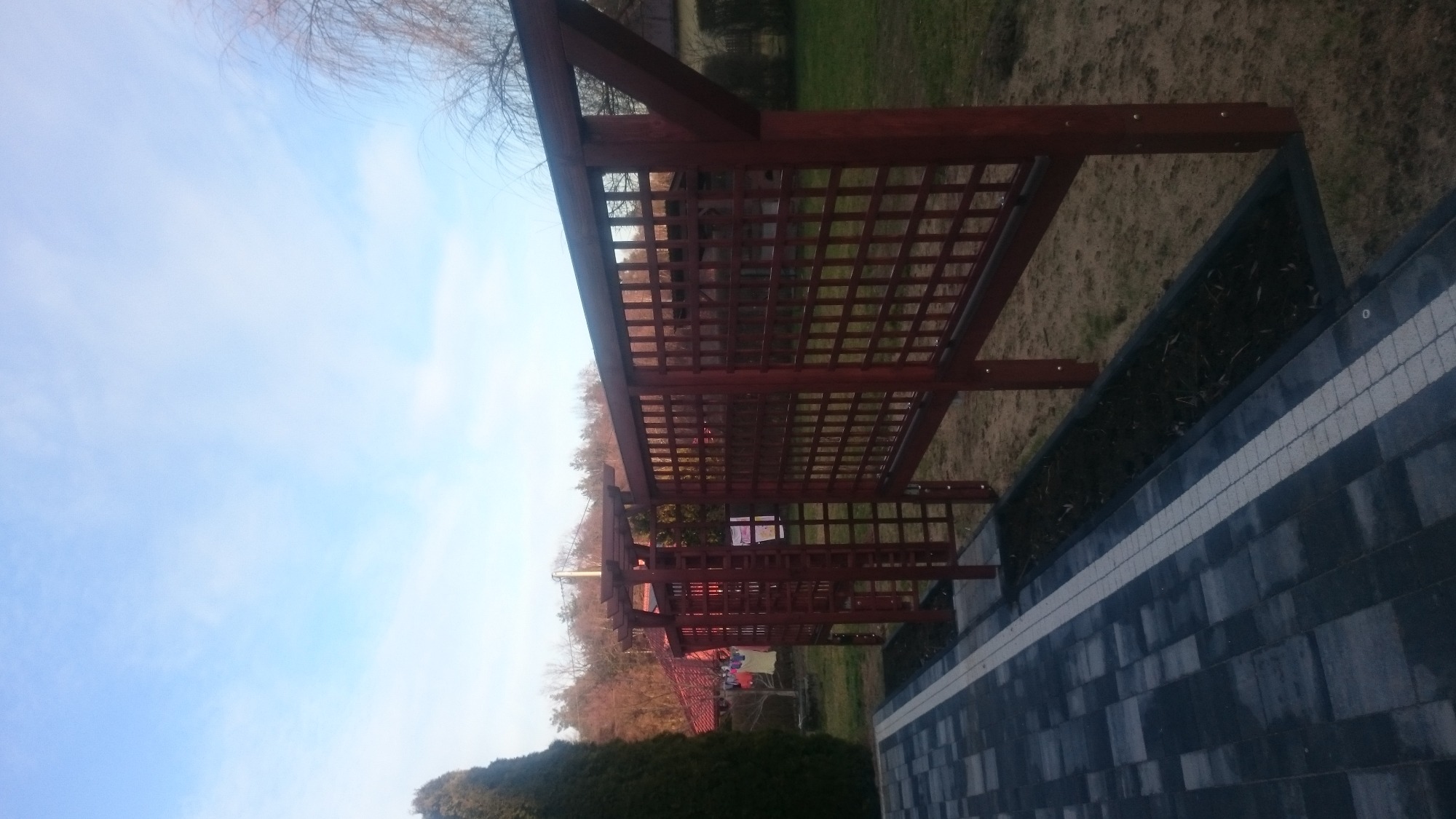 Ciemnobrązowa, drewniana pergola z kratownicą, widoczna z boku, obok wyłożonego kostką brukową chodnika i rabaty z roślinami, na tle drzew i błękitnego nieba.