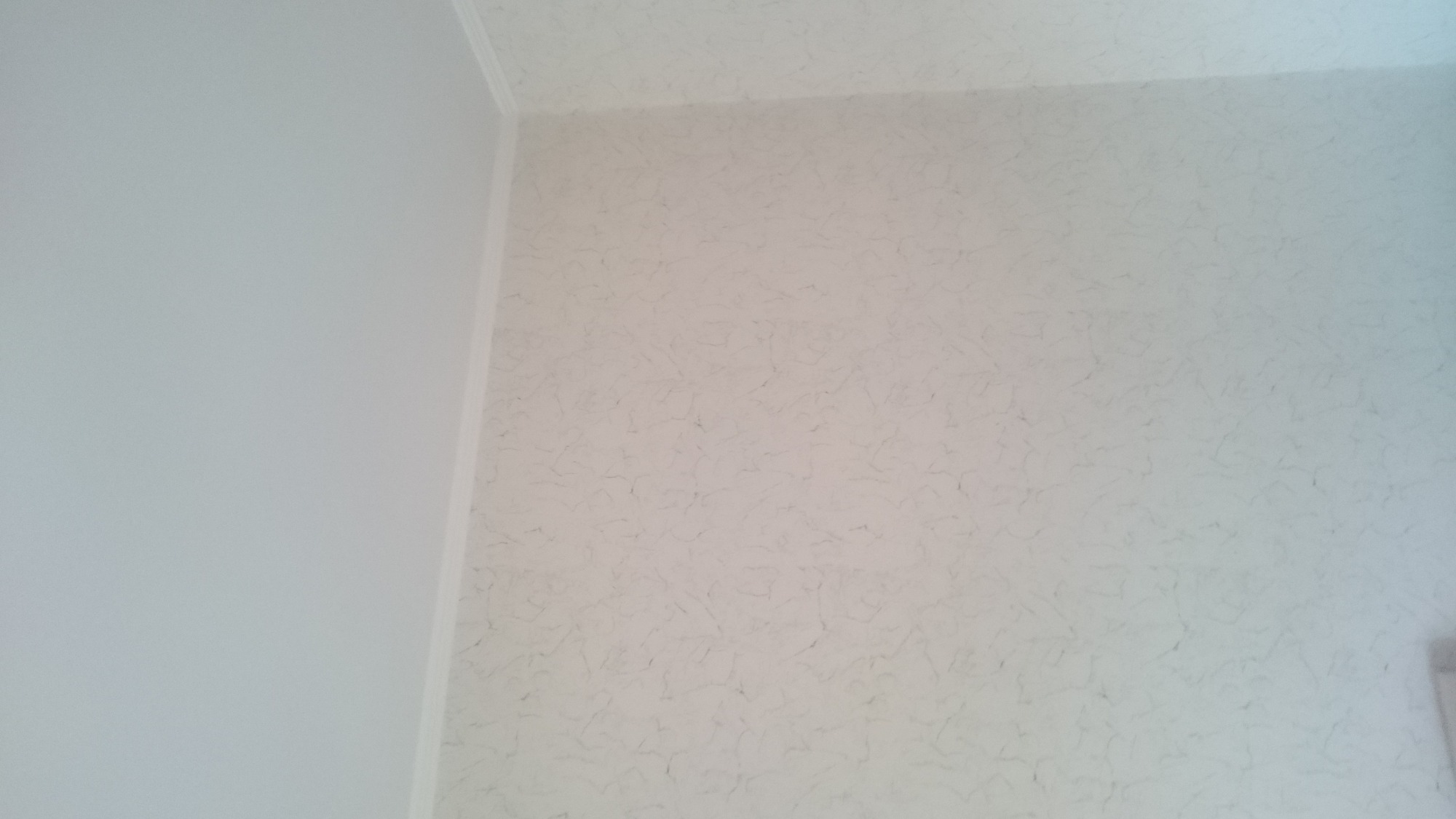 Ściana i sufit w jasnych odcieniach, pokryte tapetą o delikatnym, marmurkowym wzorze, widoczny fragment narożnika i listwy przysufitowej.