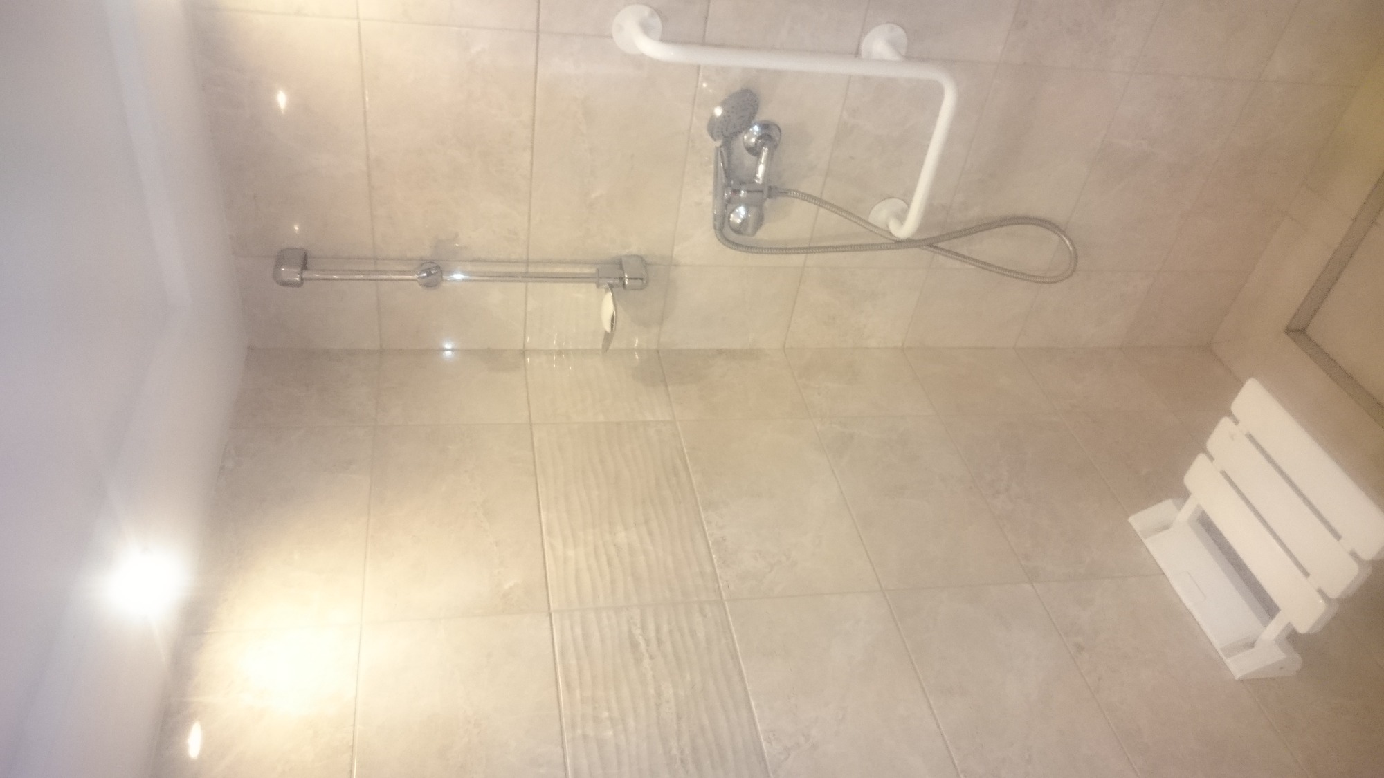 Jasna łazienka z beżowymi płytkami, zamontowany prysznic z uchwytem dla niepełnosprawnych oraz składane krzesełko prysznicowe.