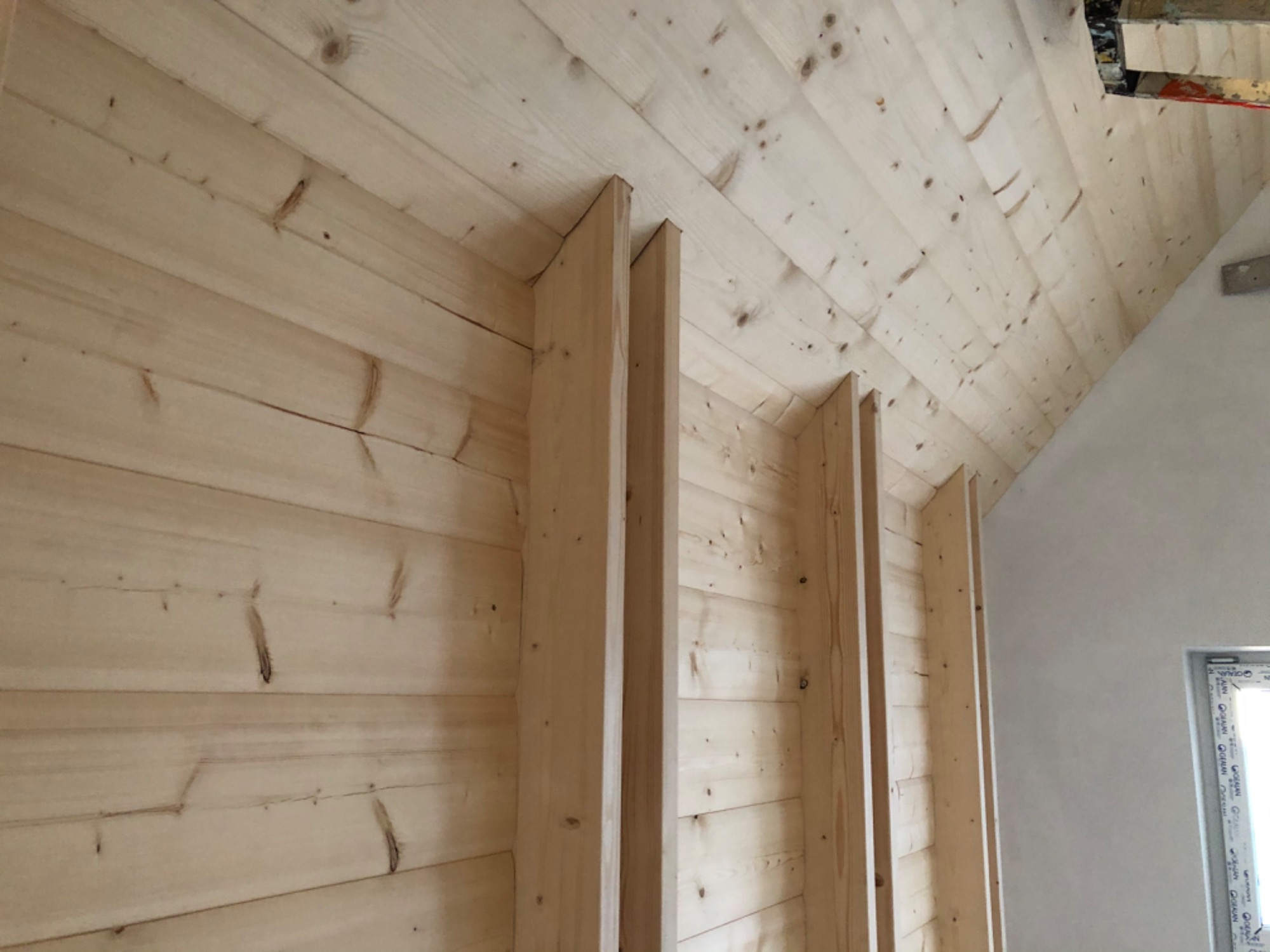 Drewniana boazeria na ścianie i suficie poddasza, widoczne pionowe listwy konstrukcyjne, fragment okna i surowa ściana w tle.