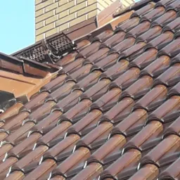 Błyszczące, brązowe dachówki ceramiczne tworzą wzór na dachu, z kominem z cegły klinkierowej w tle i błękitnym niebem u góry.