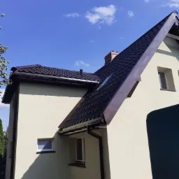 Dom jednorodzinny z brązowym dachem pokrytym dachówką, jasna elewacja, widoczne okna i rynny, błękitne niebo z chmurami w tle.