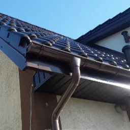 Ciemnobrązowa dachówka ceramiczna z systemem orynnowania w kolorze, zamontowana na jasnej elewacji budynku z widocznym fragmentem brązowego panelu elewacyjnego. Rynna pionowa z widocznymi złączkami.