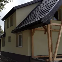 Nowa elewacja i dachówka ceramiczna na domu jednorodzinnym w Ełku, widoczne drewniane elementy konstrukcyjne werandy, teren wokół budynku w trakcie prac wykończeniowych.