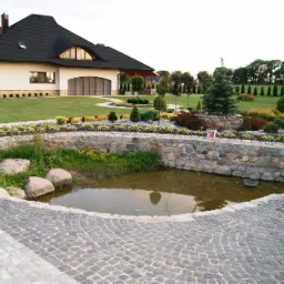Ułożona kostka granitowa wokół oczka wodnego w ogrodzie z widokiem na dom z czarnym dachem i zielonym trawnikiem.
