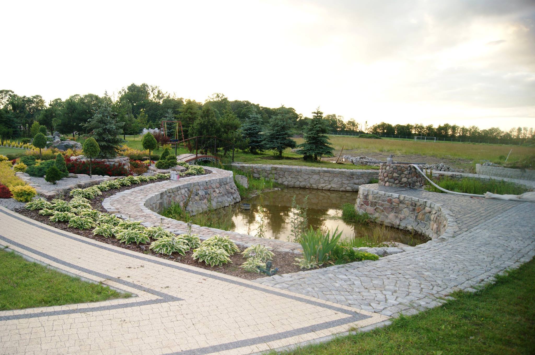 Ogród z oczkiem wodnym otoczonym kamienną obudową i ścieżką z kostki brukowej, z widocznymi elementami krajobrazu w tle.