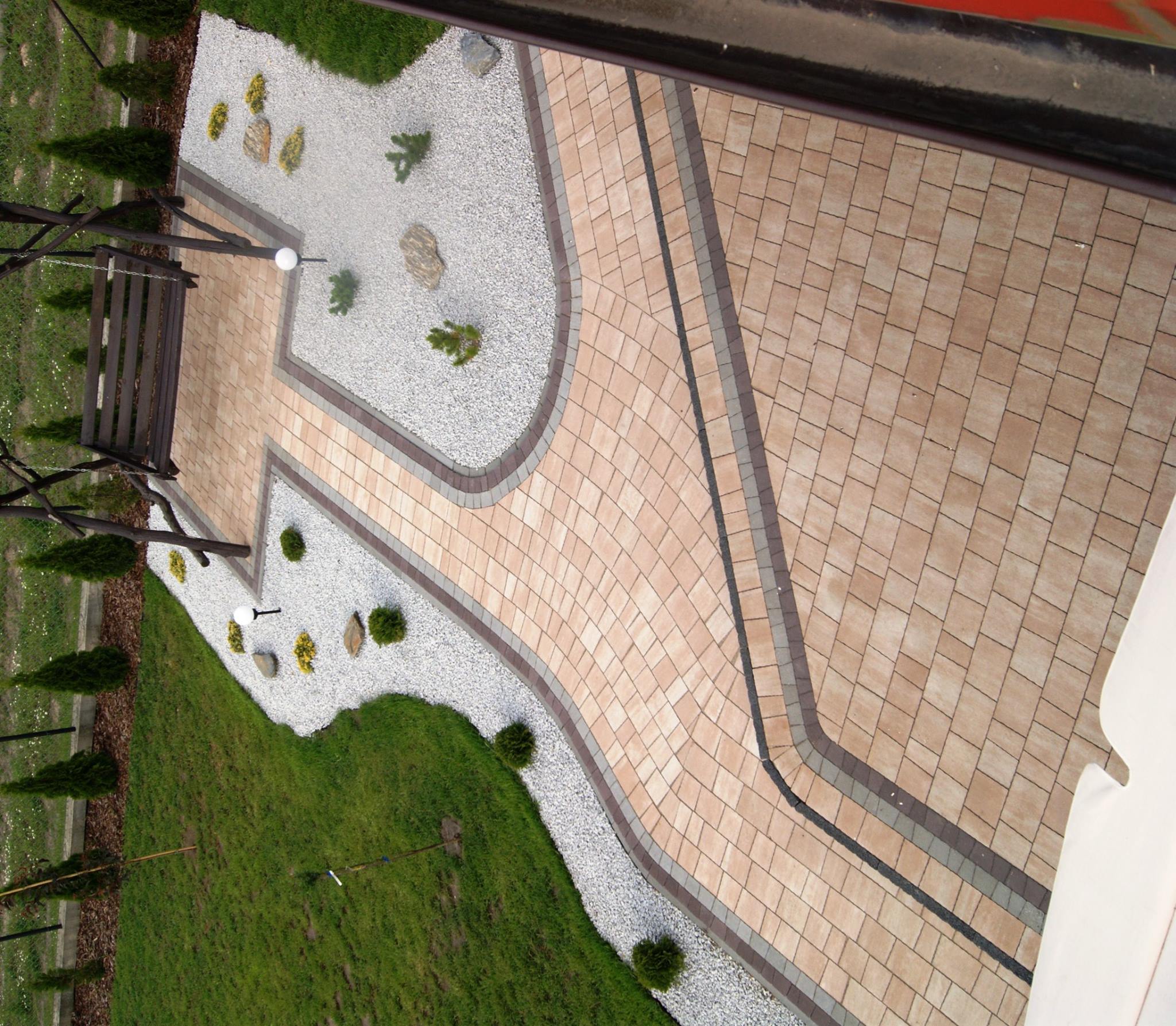 Widok z góry na ścieżkę z beżowej kostki brukowej, obrzeża z szarej kostki, biały żwir, trawnik i małe drzewka iglaste.