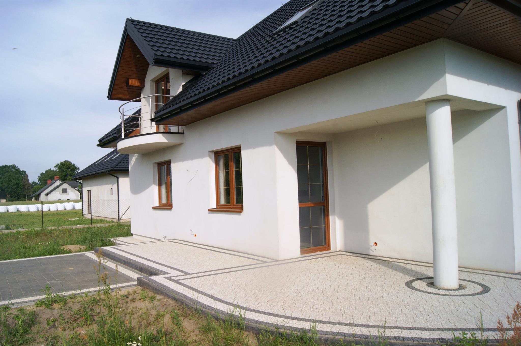 Nowoczesny dom jednorodzinny w Ołdakach z jasną elewacją i ciemną dachówką, z wyłożonym kostką brukową tarasem i podjazdem. Widoczne detale wykończeniowe, takie jak obramowania okien i balustrada...