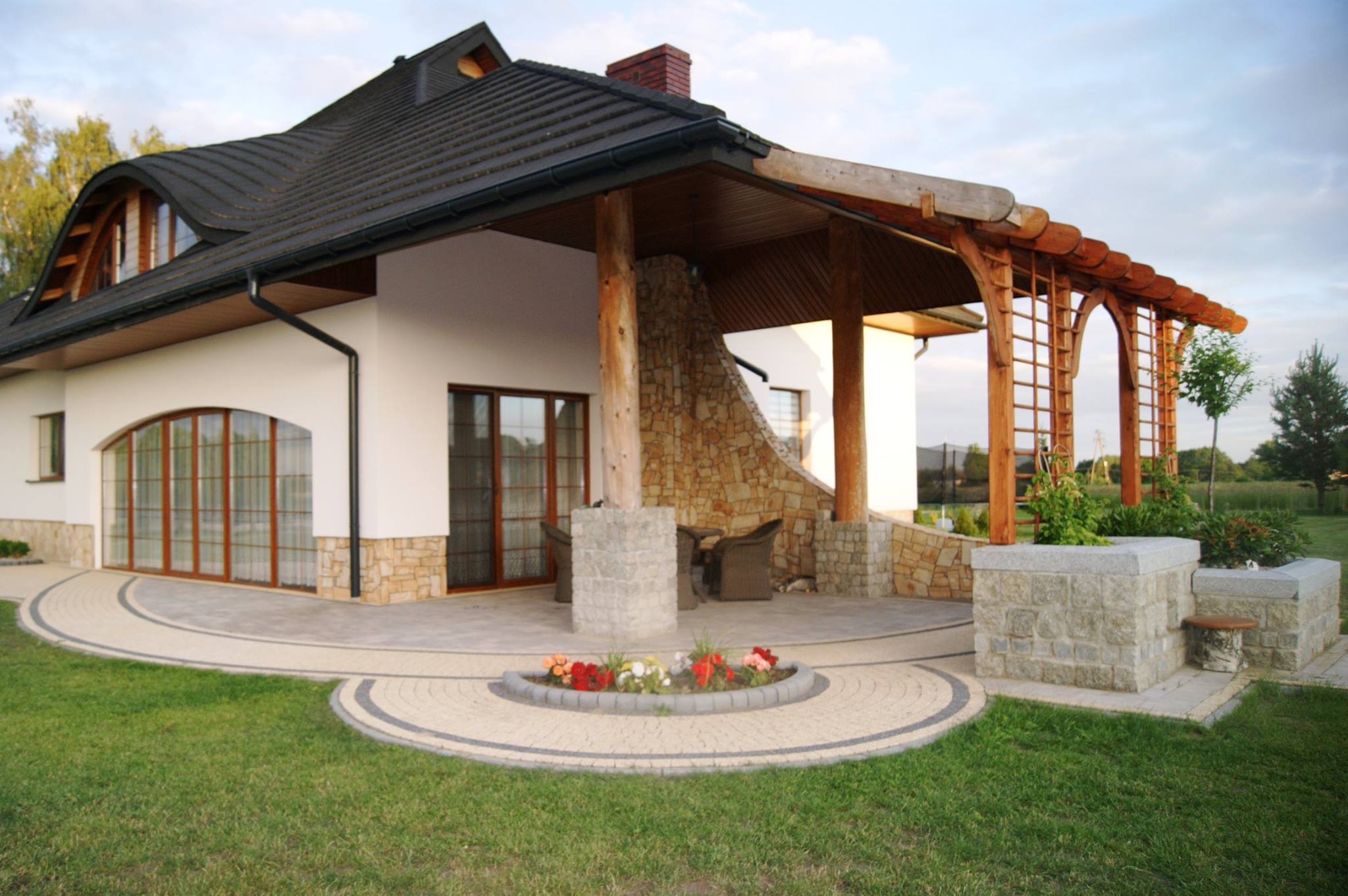 Elegancki dom z tarasem wyłożonym jasną kostką brukową w okrągły wzór, ozdobiony rabatą kwiatową i kamienną pergolą, widok na zadbany trawnik.