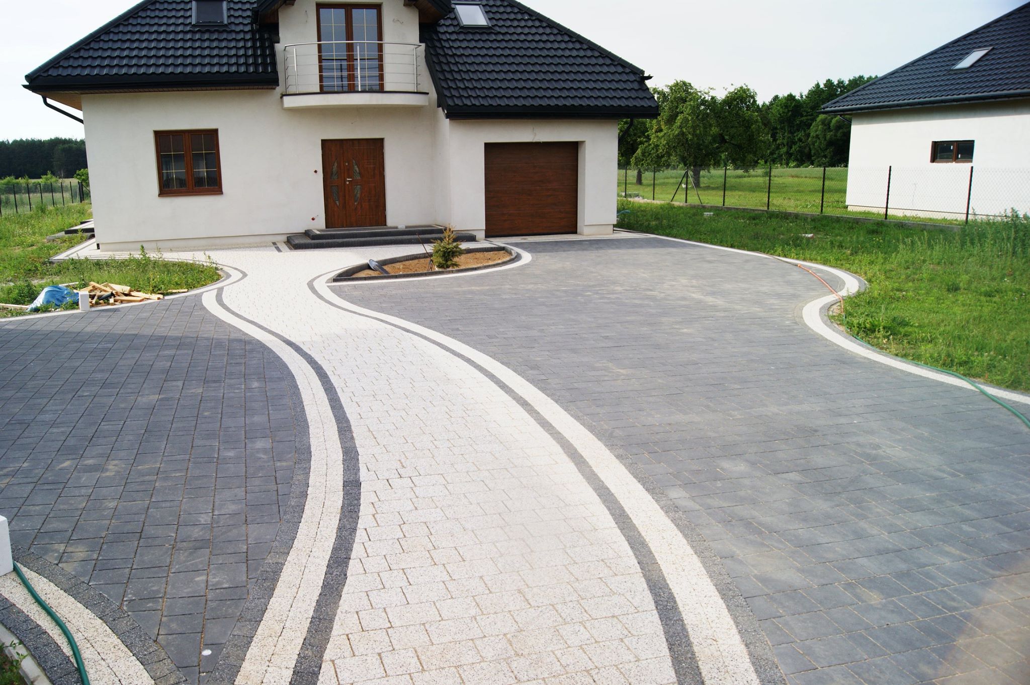 Ułożona kostka brukowa w dwóch odcieniach szarości i bieli tworząca falisty wzór podjazdu przed nowoczesnym domem z brązową stolarką okienną i drzwiową.