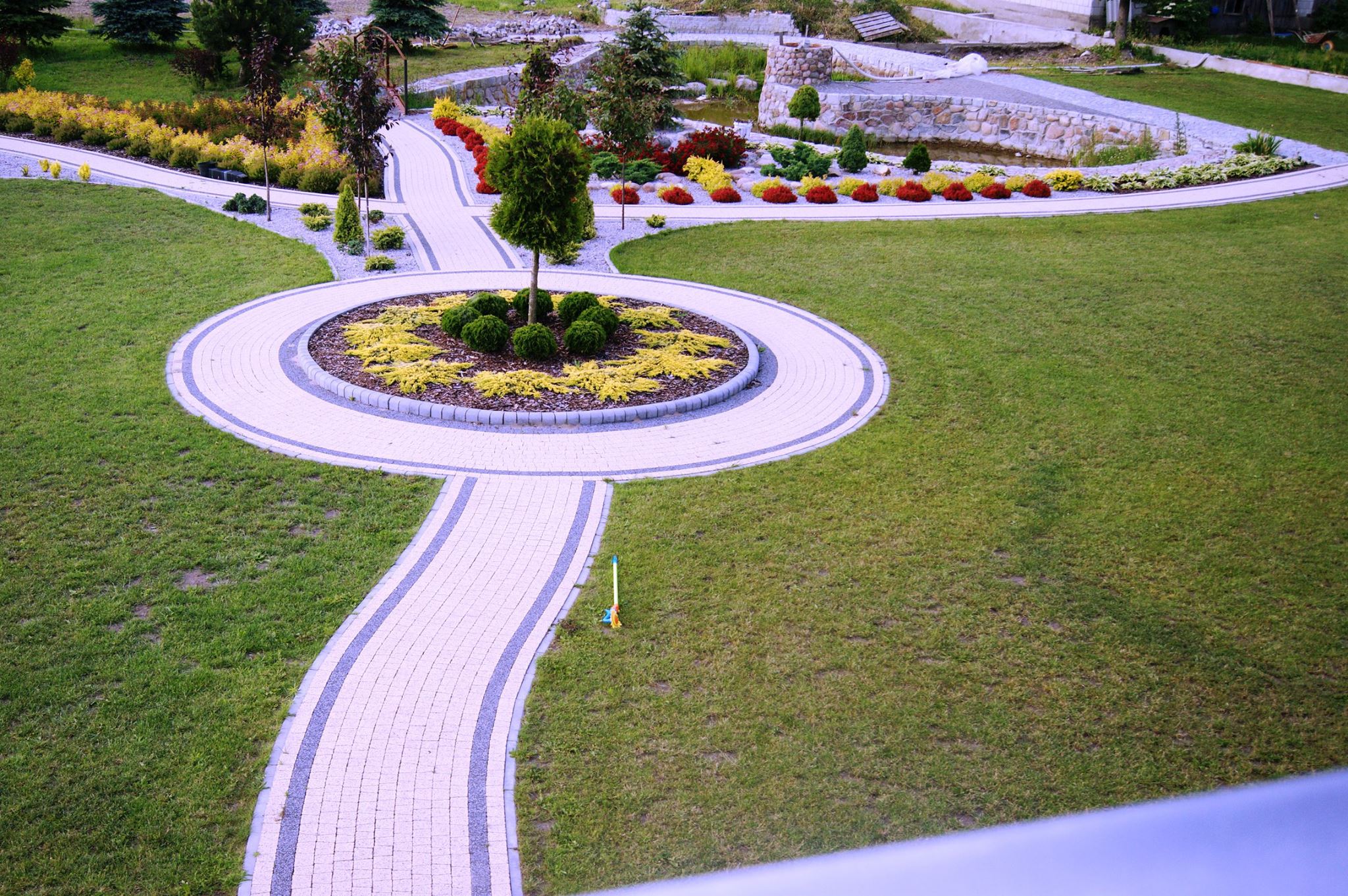 Widok z góry na ścieżki z kostki brukowej w ogrodzie z okrągłym klombem i stawem w tle.