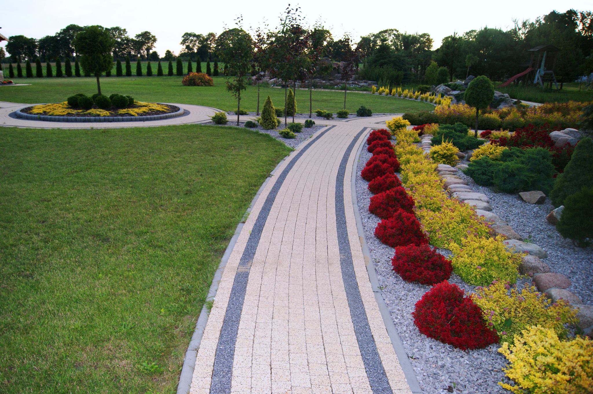 Kremowa ścieżka z kostki brukowej z szarymi pasami, otoczona czerwonymi i żółtymi krzewami, prowadząca przez zadbany ogród z trawnikiem i drzewami.