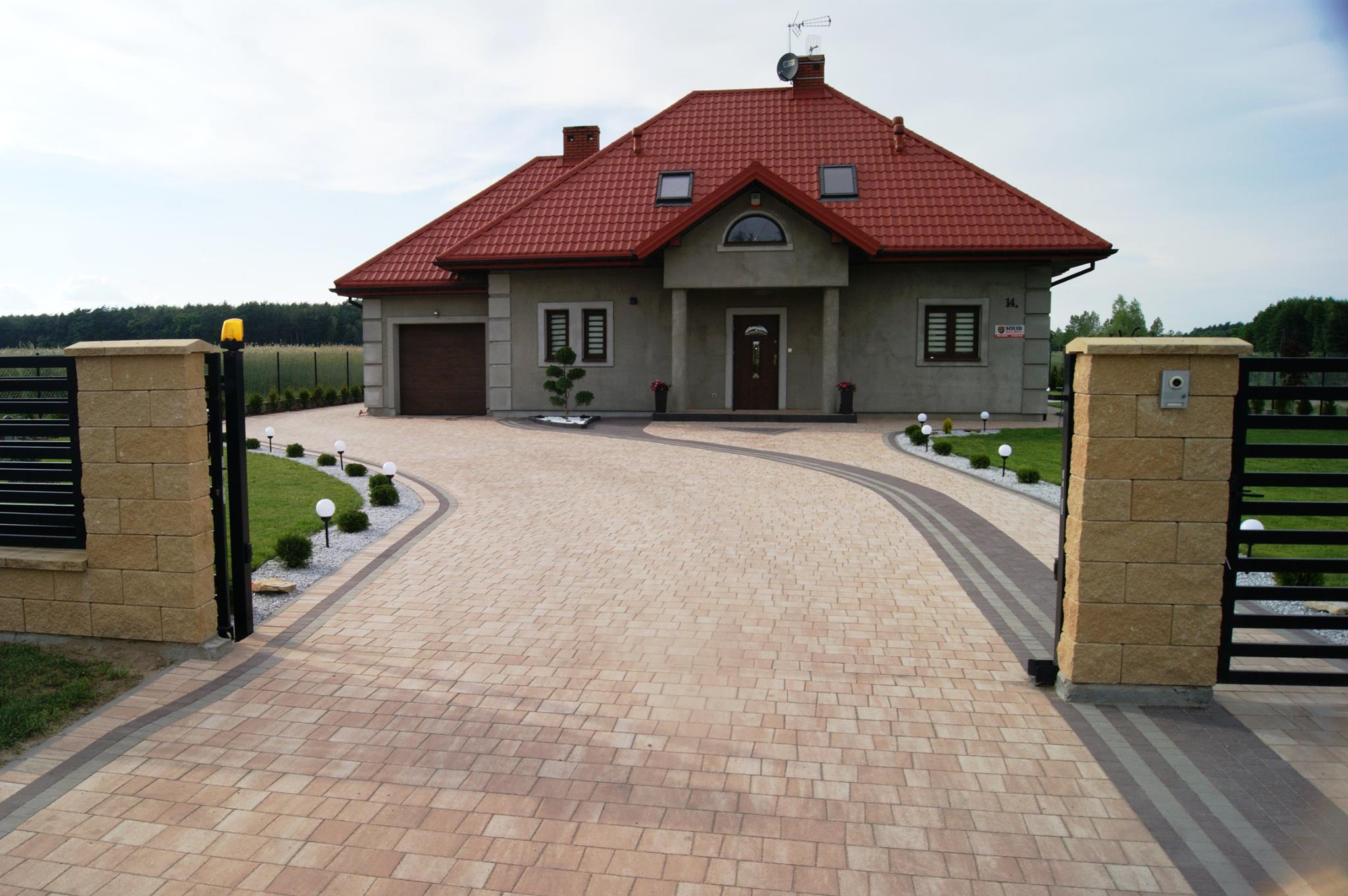 Ułożona kostka brukowa w kolorze beżowym tworząca podjazd do domu z czerwonym dachem, widoczne ogrodzenie z kamiennymi słupkami i oświetlenie wzdłuż ścieżki.