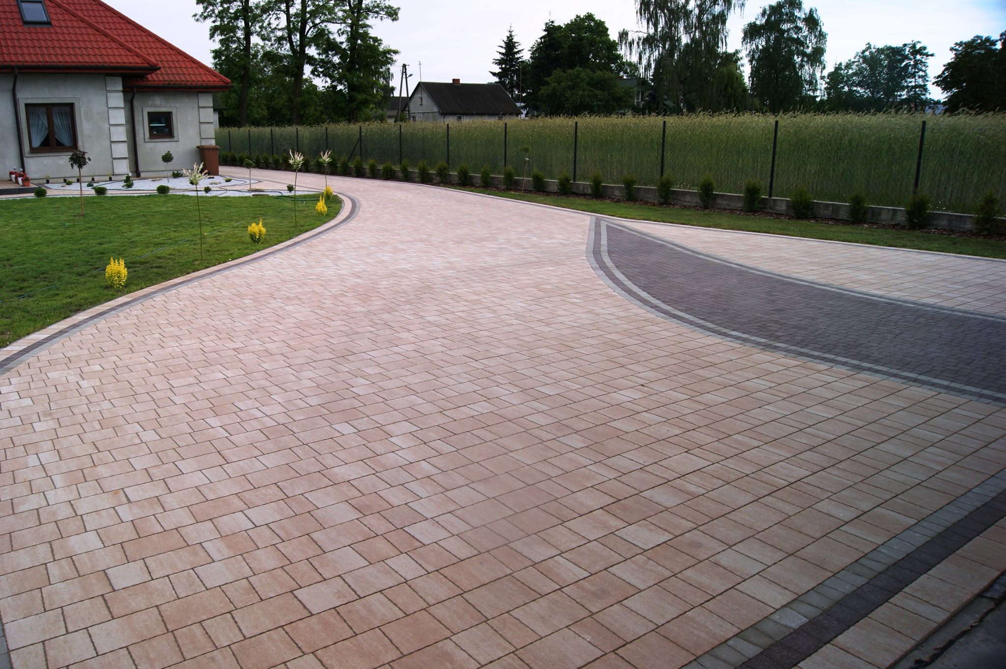 Zakrzywiony podjazd z beżowej kostki brukowej prowadzący do domu z czerwonym dachem, obrzeża z ciemniejszej kostki, trawnik z niskimi krzewami i wysoką trawą w tle.