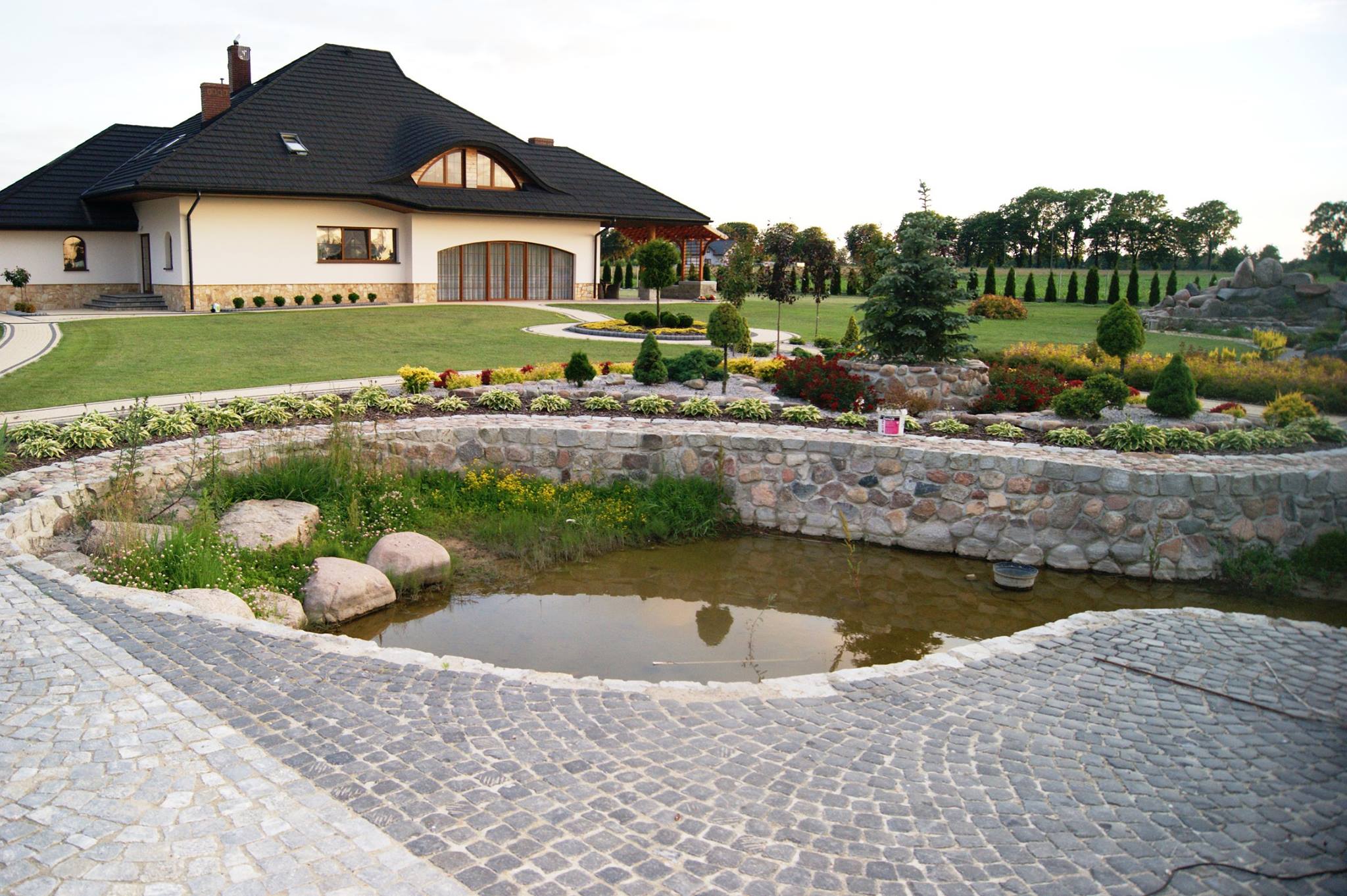 Ułożona kostka granitowa wokół oczka wodnego w ogrodzie z widokiem na dom z czarnym dachem i zielonym trawnikiem.