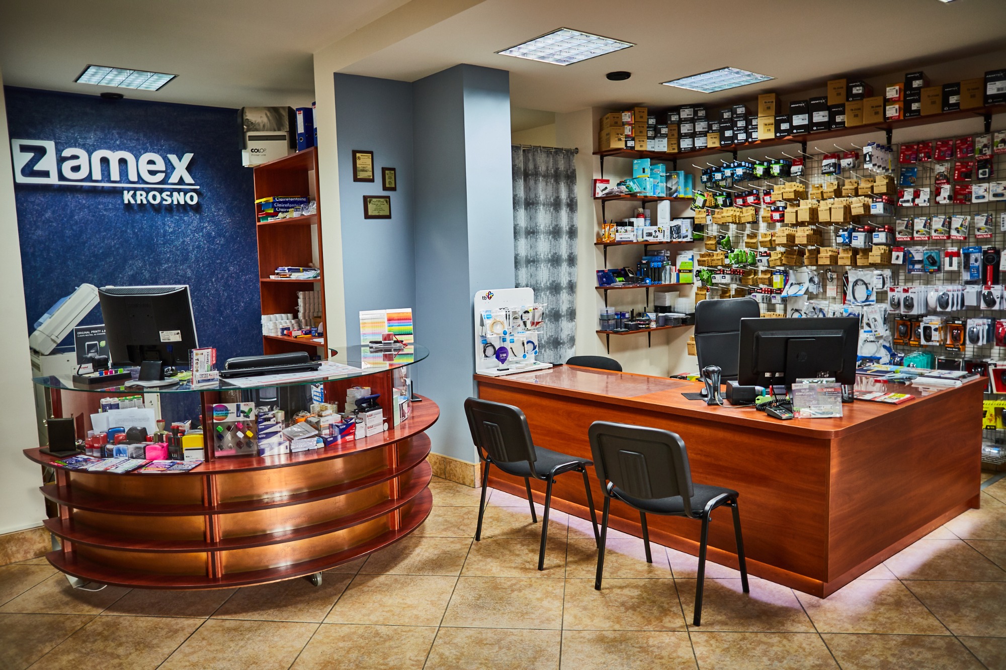 Wnętrze sklepu komputerowego Zamex w Krośnie, z ladą recepcyjną, półkami pełnymi akcesoriów i logo firmy na ścianie.