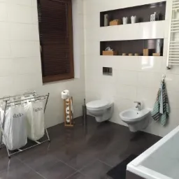 Nowoczesna łazienka z brązowymi płytkami podłogowymi, białymi płytkami ściennymi, wiszącą toaletą i bidetem, drewnianymi żaluzjami w oknie oraz wnękami w ścianie z dekoracjami.